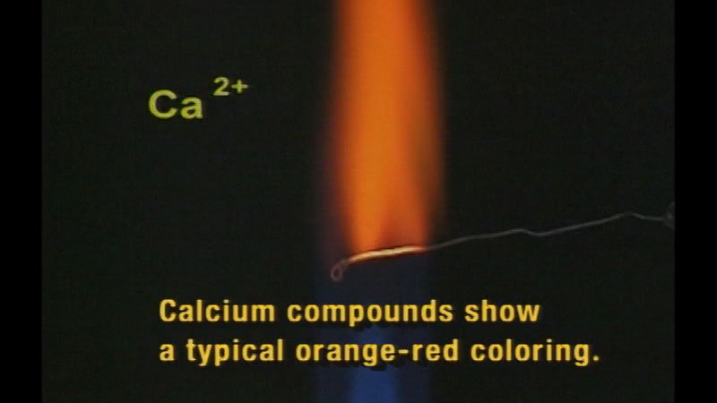 Open flame with a piece of metal in it. Flame below the metal is blue, above the metal is orange. Ca2+. Caption: Calcium compounds show a typical orange-red coloring.
