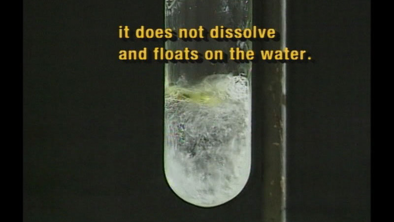 Test tube with a clear substance on the bottom. At the top of the substance is a light-yellow patch. Caption: it does not dissolve and floats on the water.