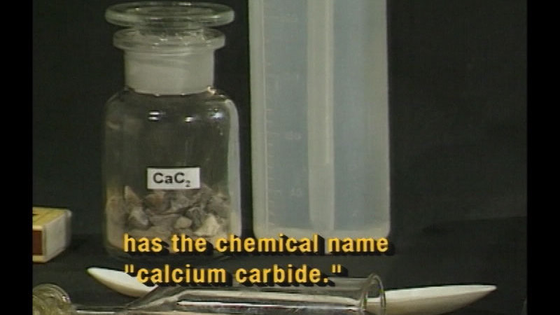 Glass container labelled as CaC2 with a gray, rocky substance sitting among other scientific instruments. Caption: has the chemical name "calcium carbide."