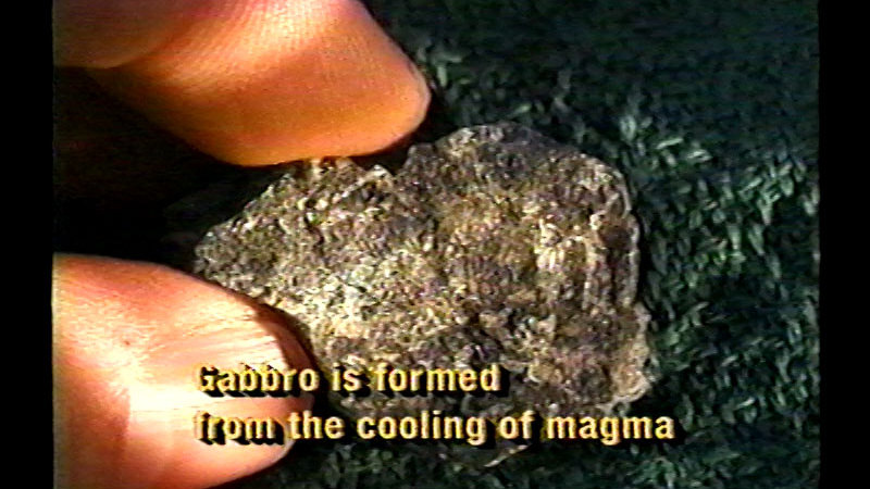 Closeup of fingers picking up a sliver of grayish rock. Caption: Gabbro is formed from the cooling of magma.
