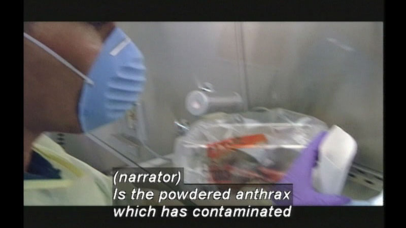 Person wearing a protective gown, mask and gloves handling materials. Caption: (narrator) Is the powdered anthrax which has contaminated