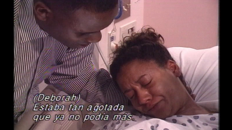 A woman in a hospital setting in labor with someone next to her supporting her neck. Spanish captions.