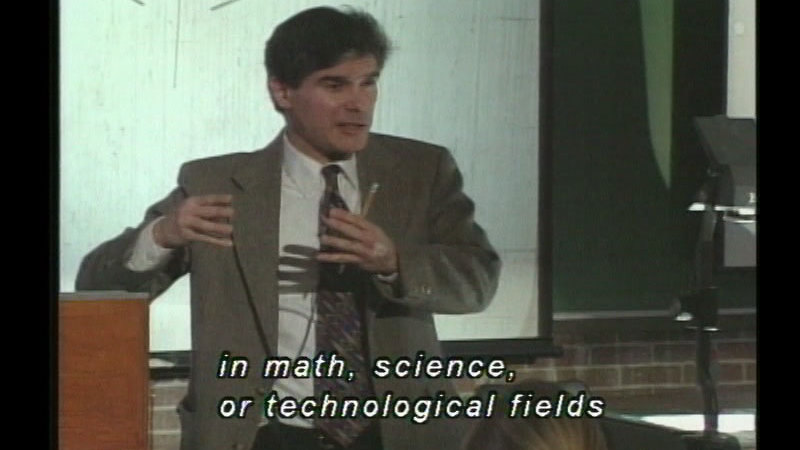 Person speaking in front of a projector screen. Caption: in math, science, or technological fields.