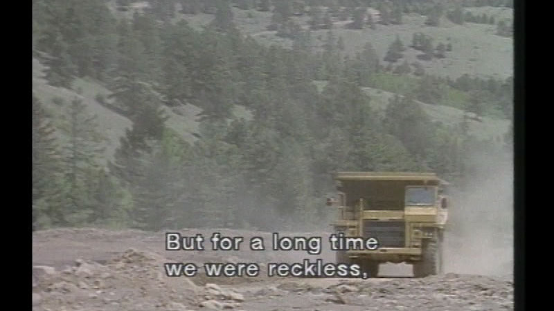 Industrial machinery leaving a cloud of dust in its wake, driving across a bare rocky landscape with a partially forested mountain in the background. Caption: But for a long time we were reckless,