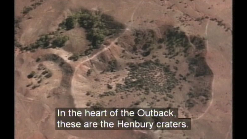 Aerial view of a large depression in the earth. Caption: In the heart of the Outback, these are the Henbury craters.
