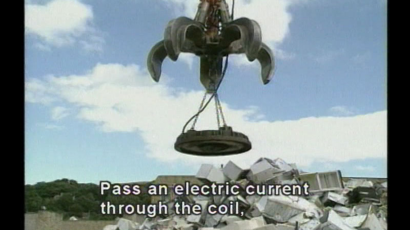 Industrial claw with giant magnet descending on a pile of trash. Caption: Pass an electric current through the coil,