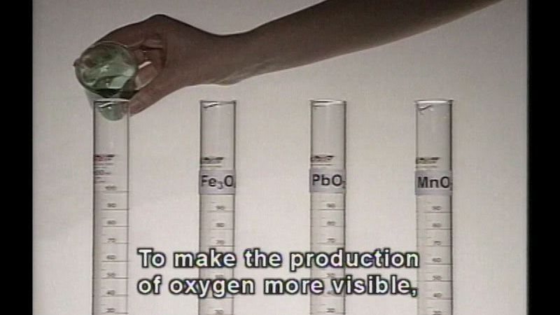 Four test tubes. Three are labelled as Fe3O2, PbO2, and MnO2. The fourth is unlabeled and a beaker of colored liquid is being poured into it. Caption: To make the production of oxygen more visible,