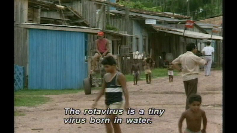 People walking on a dirt path. Buildings with corrugated metal line the road. Caption: The rotavirus is a tiny virus born in water.