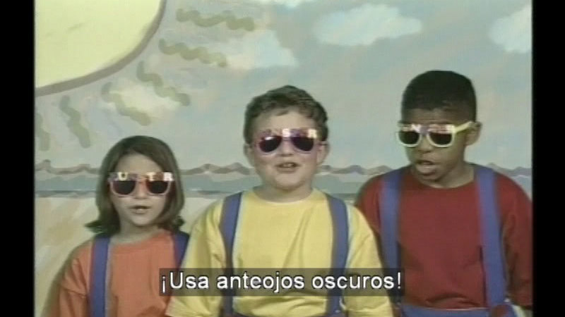 Children wearing dark sunglasses against a backdrop of the beach. Spanish captions.