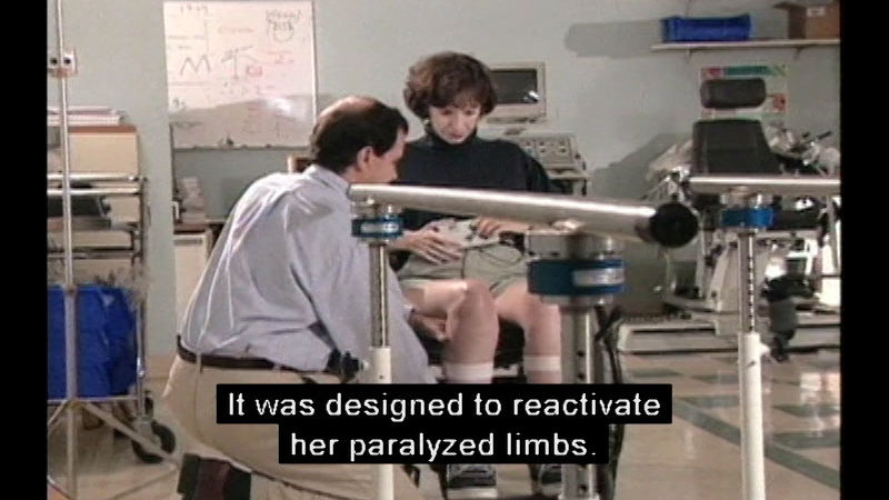 Woman in a wheelchair with straps around her legs and a control device in her hands while someone crouches in front of her in a lab setting. Caption: It was designed to reactivate her paralyzed limbs.