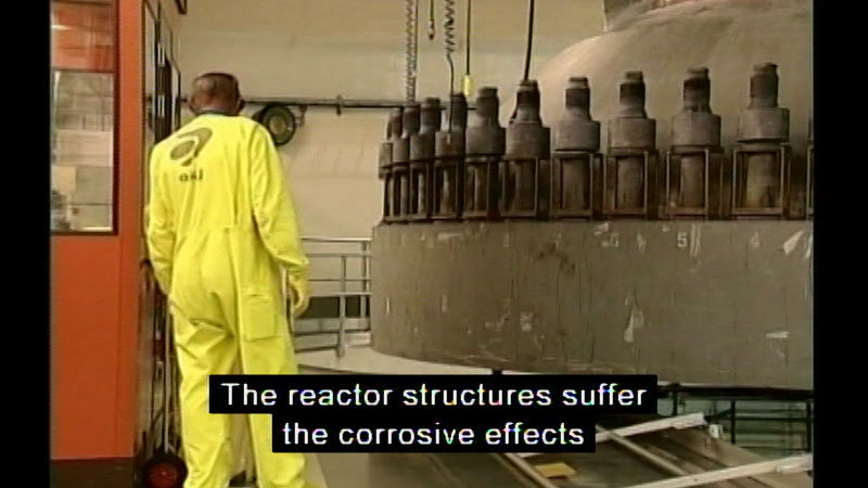 Person in a yellow jumpsuit standing next to a large piece of industrial equipment. Caption: The reactor structures suffer the corrosive effects