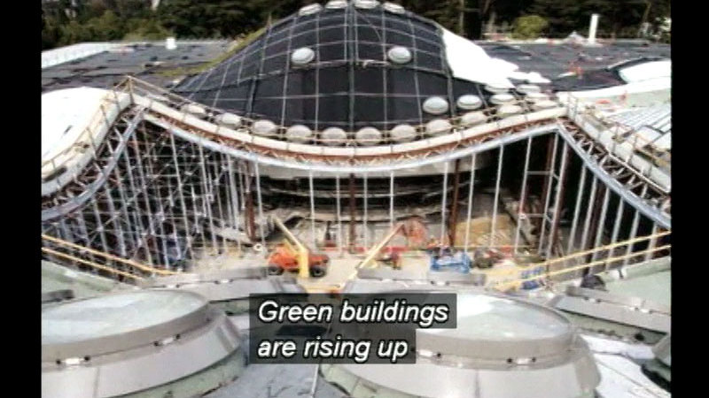 A building with a curving roofline still under construction. Caption: Green buildings are rising up
