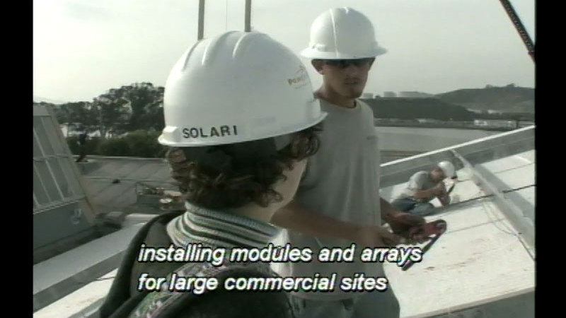 People on a rooftop wearing hard hats with tools in their hands. One hard hat says "Solari" on the back. Caption: installing modules and arrays for large commercial sites