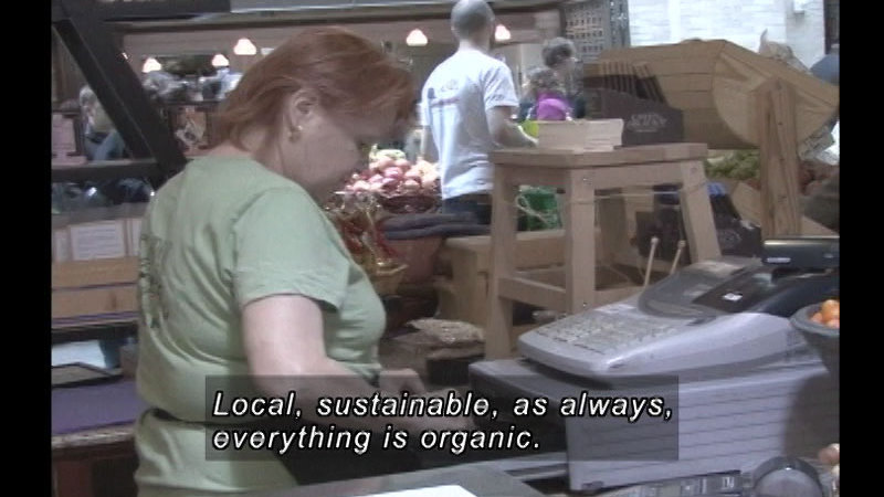 Person at a cash register in a store filled with people and produce. Caption: Local, sustainable, as always, everything is organic.