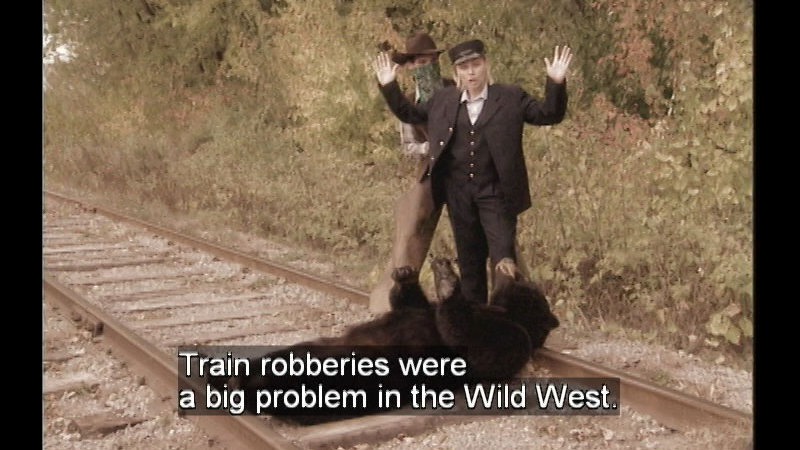 A bear laying across train tracks with two people standing above it. Caption: Train robberies were a big problem in the Wild West. 