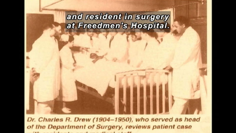 Black and white photograph of a large group of doctors standing around patient in a hospital bed. Dr. Charles R. Drew (1904-1950) who served as head of the Department of Surgery, reviews patient case Caption: and resident in surgery at Freedmen's Hospital.