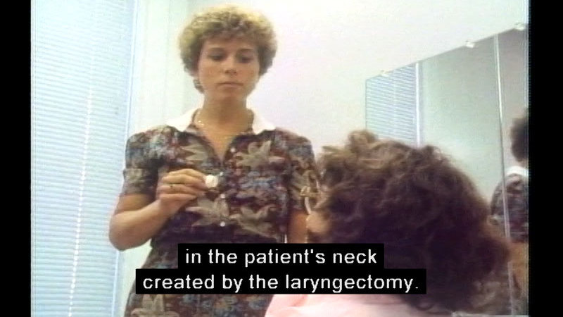 Person standing and holding a small object in their hand while facing a person who is sitting. Caption: in the patient's neck created by the laryngectomy.