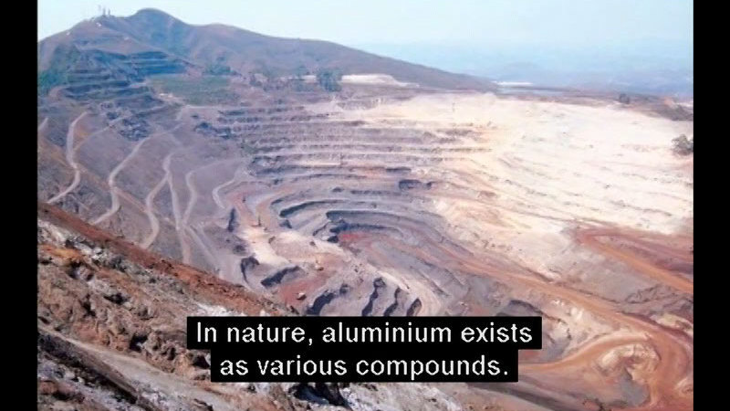Terraced sloping sides lead to a deep depression in a hillside of brown, gray, and red dirt. Caption: In nature, aluminum exists as various compounds.