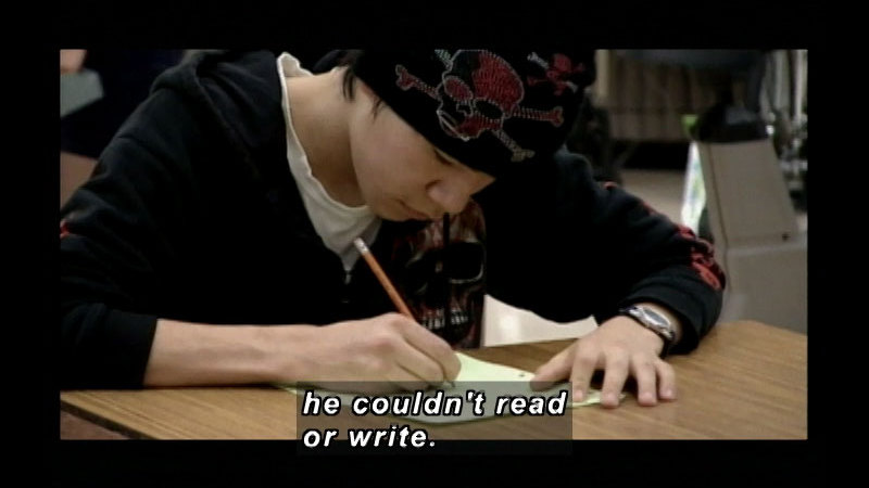 A teenager wearing a beanie with skull and crossbones and a hoodie with a skull, sitting at a desk and writing on a piece of paper. Caption: he couldn't read or write.