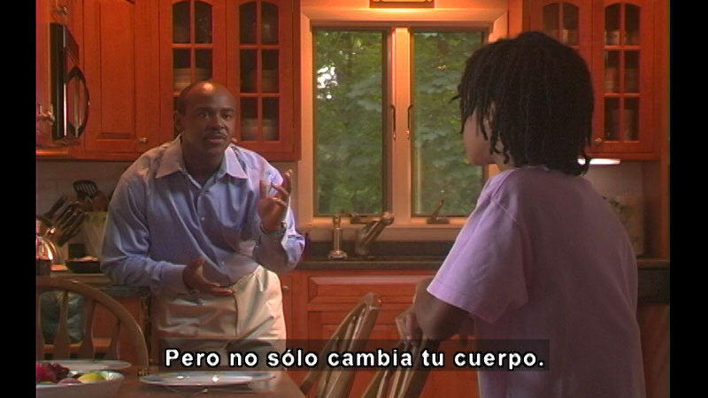Adult and young person talking in a kitchen. Spanish captions.