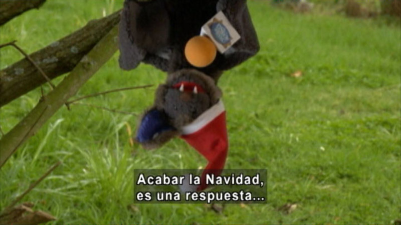 Puppet of a bat handing upside down holding a microphone and wearing a Santa hat. Spanish captions.