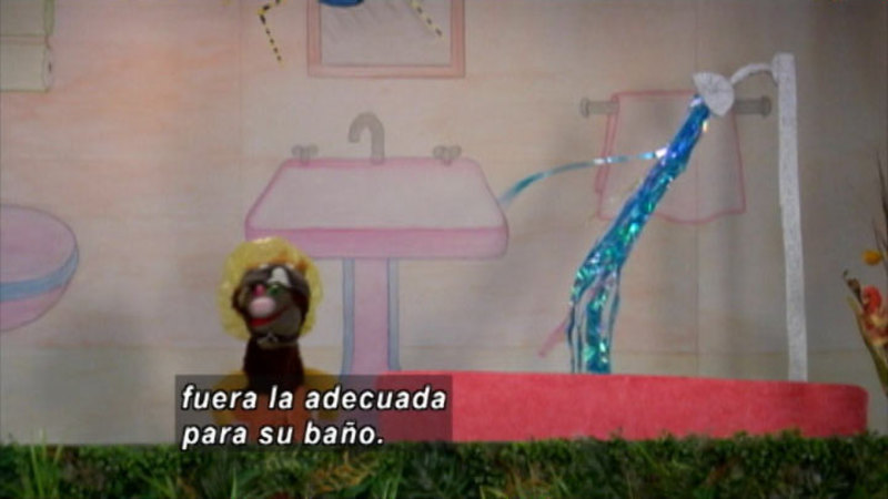 A puppet standing in a staged bathroom. Spanish captions.