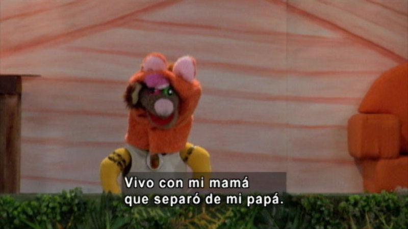Animal puppet in front of a building. Spanish captions.