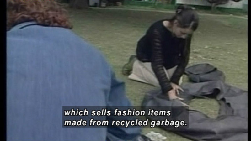 Person kneeling on the ground next to a pile of dark material. Caption: which sells fashion items made from recycled garbage.