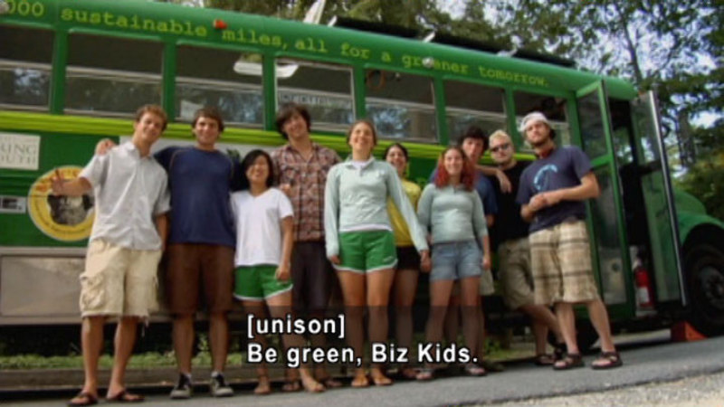 10 people standing closely together in front of a school bus painted green. Caption: [unison] Be green, Biz Kids.