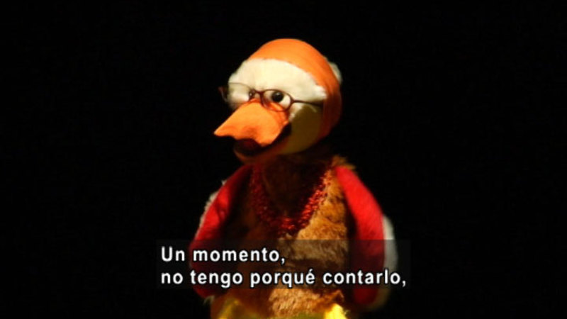 A bird puppet wearing glasses. Spanish captions.