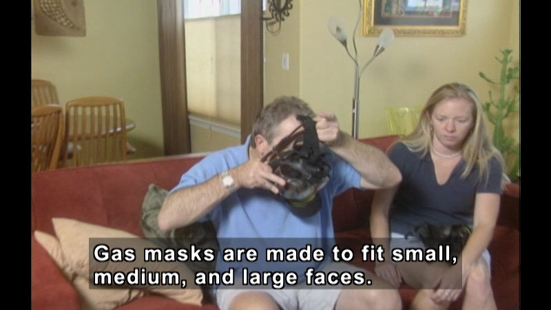 Two people sit on a couch. One is putting on a gas mask. Caption: Gas masks are made to fit small, medium, and large faces.