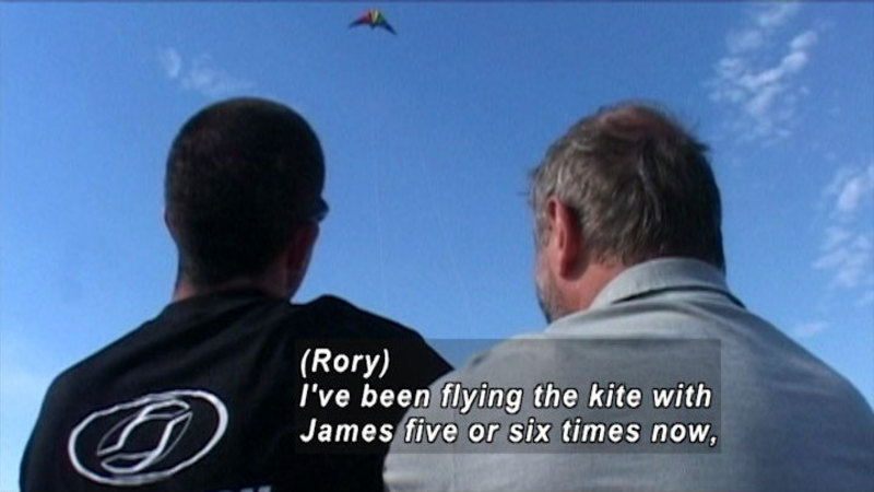 Two people looking up at a kite flying in the sky. Caption: {Rory) I've been flying the kite with James five or six times now,