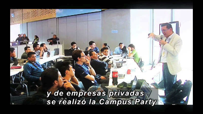 Teacher speaking in front of a lecture hall full of students. Spanish captions.