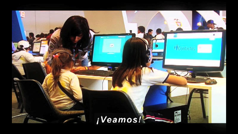 Teacher leaning down to help a student at a computer. Spanish captions.