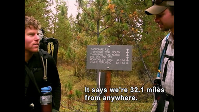 Two people in hiking gear standing next to a sign showing destinations and distance. Caption: It says we're 32.1 miles from anywhere.