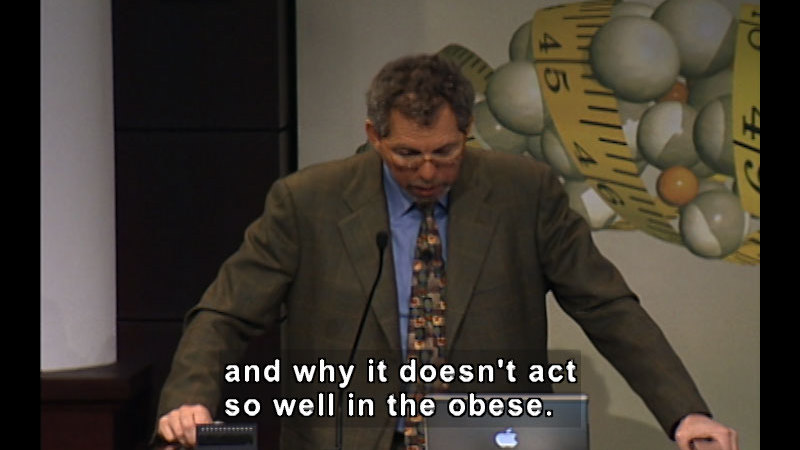 Person Speaking from a podium. Caption: and why it doesn't act so well in the obese.