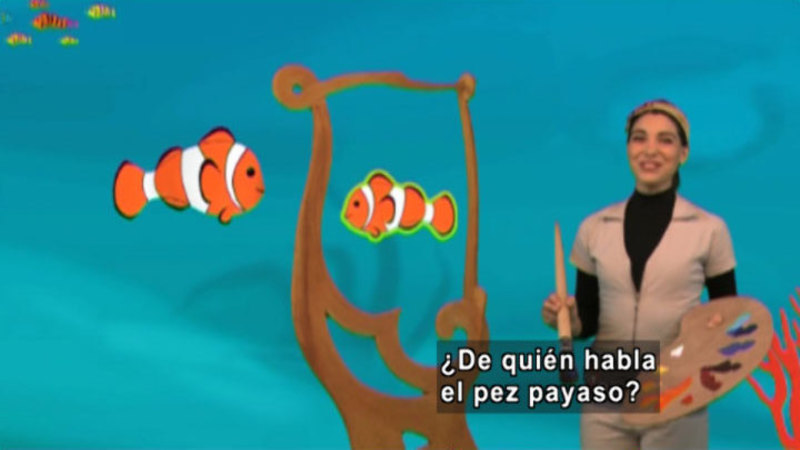 A woman with a painting pallet and a paintbrush. On her painting there are two clown fish. Spanish captions.