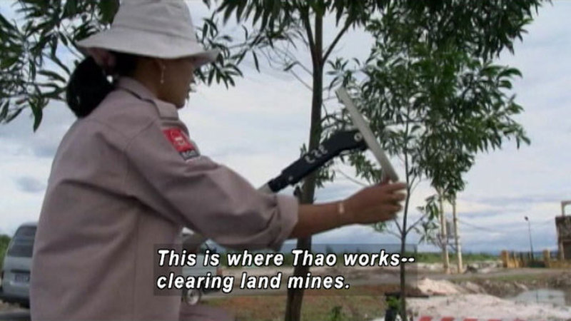 Person wearing khaki hat and shirt holding a tool. Low building and vehicle in background. Caption: this is where Thao works -- clearing land mines.