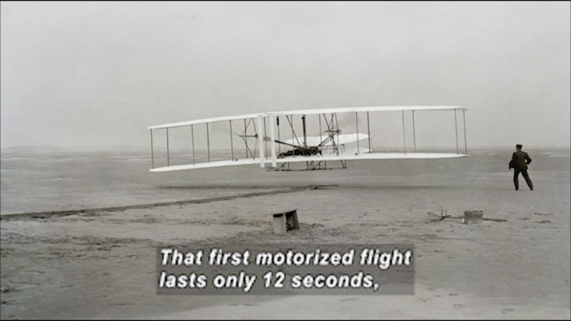 Black and white photograph of an early plane with a person standing next to it. Caption: That first motorized flight lasts only 12 seconds,