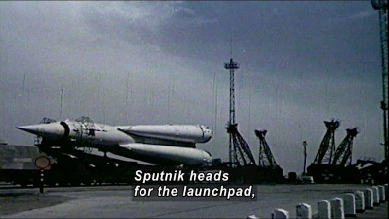 Black and white photograph of a rocket-like object on its side in an industrial area. Caption: Sputnik heads for the launchpad,