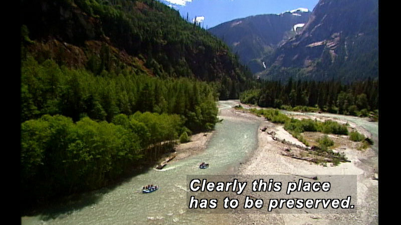 Tall snowcapped mountains covered in lush green trees and a light green river with two rafts winding through the valley. Caption: Clearly this place has to be preserved.