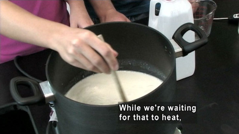 Person stirring a white liquid in a large pot. Caption: While we're waiting for that to heat,