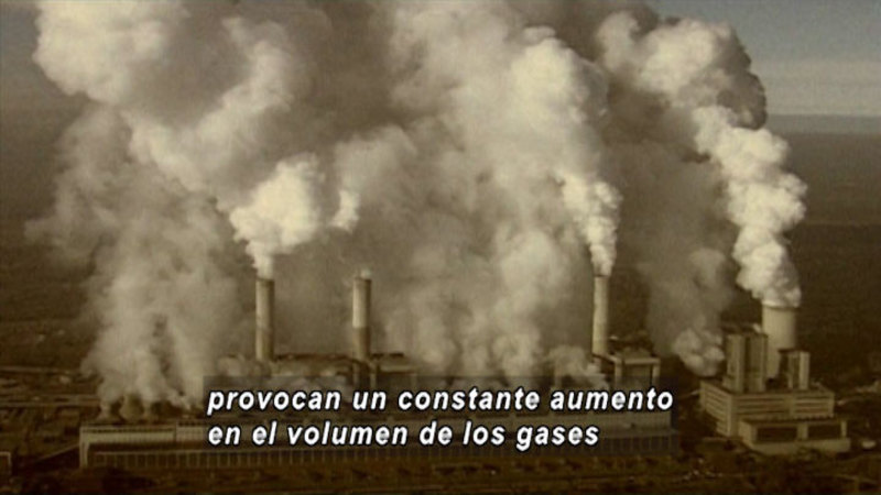 An industrial plant with multiple smokestacks emitting pollutants. Spanish captions.