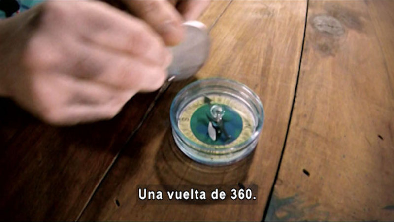 Person holding an object near a compass. The arrow on the compass points towards the object. Spanish captions.