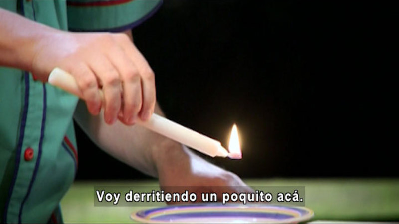 Person with a lit candle melting the wax into a dish. Spanish captions.