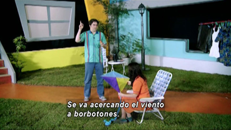 One person stands while holding a kite string and the second person sits in a chair while holding the kite. Spanish captions.