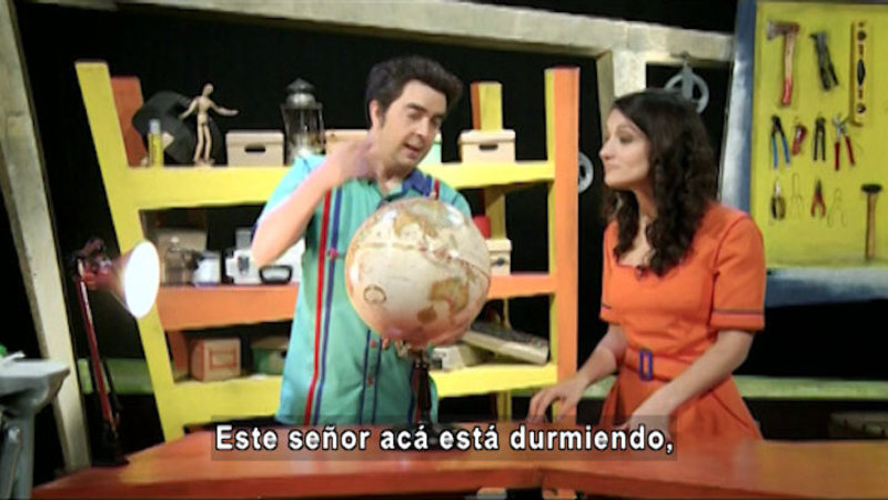 Two people standing while one gestures at a globe. Spanish captions.