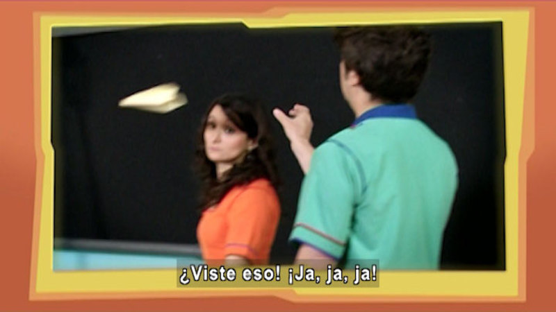 Two people standing while a paper airplane flies through the air. Spanish captions.