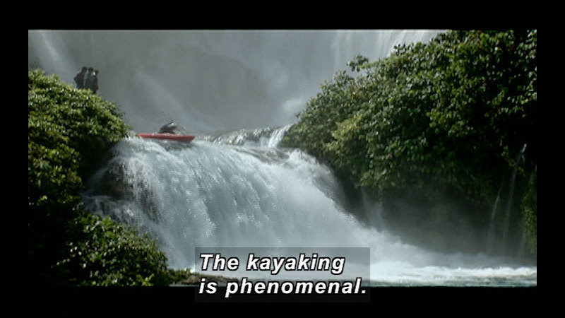 A kayak is about to go over a small waterfall on a river lined by lush greenery. Caption: The kayaking is phenomenal.