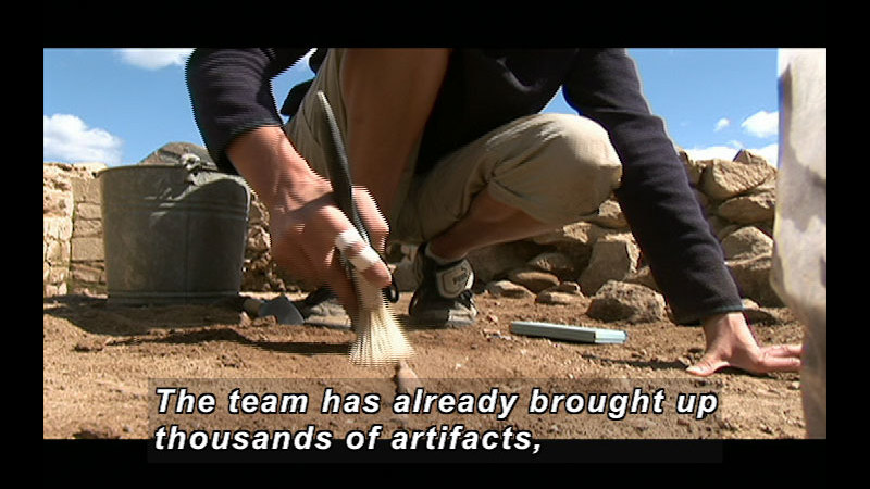 Person crouching and using a brush to expose an item in the dirt. Caption: The team has already brought up thousands of artifacts,